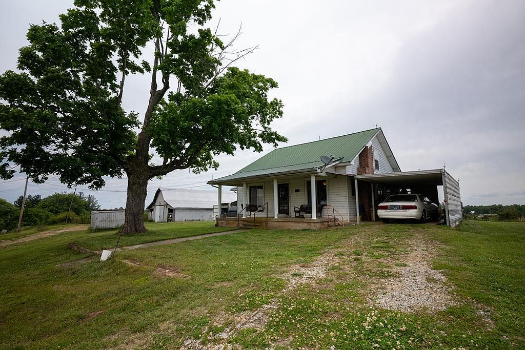 The 1955 farmhouse from the side, under a mature shade tree with the green metal roof, white clapboard siding, and the carport beyond