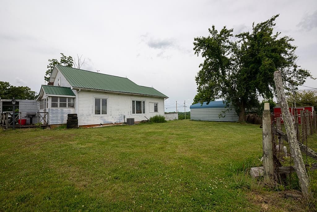 Backyard view showing the rear of the farmhouse, a blue metal outbuilding, and a wood fenceline at the edge of the pasture
